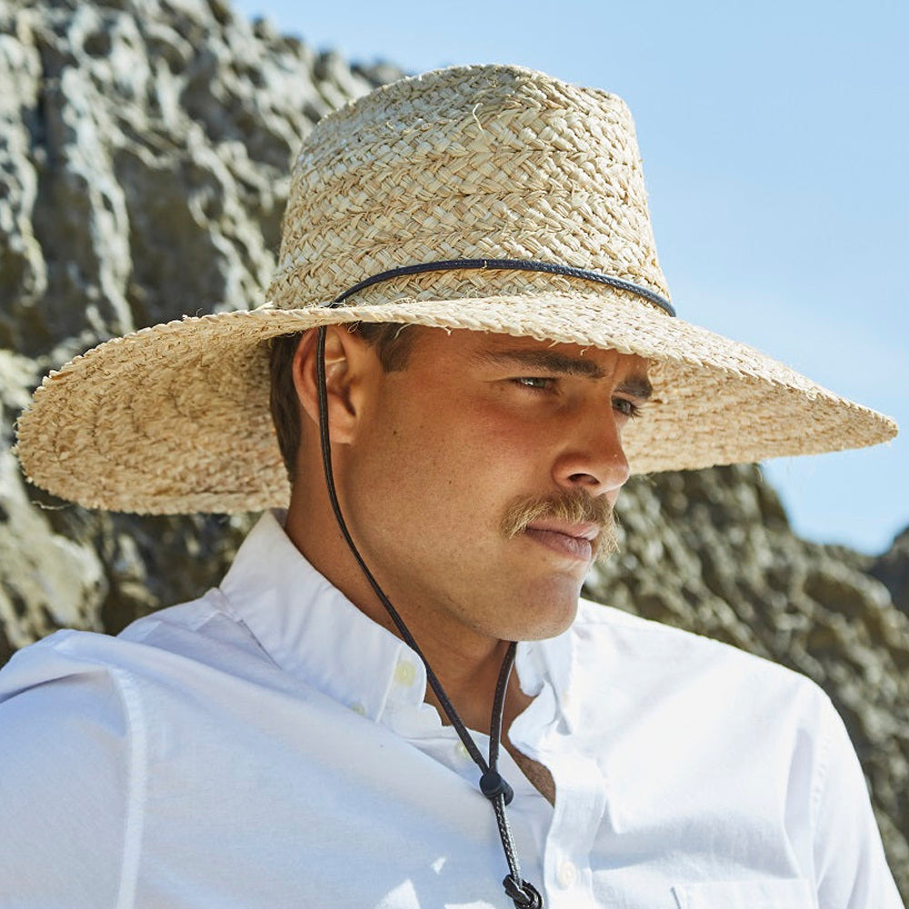 Man wearing a natural colored straw hat  and white shirt standing against a cliffside background