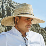 Man wearing a natural colored straw hat  and white shirt standing against a cliffside background