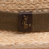 Close-up of a natural colored straw hat with a brown band and logo, featuring a golden colored golf club bag and golf ball ball pin on top of it