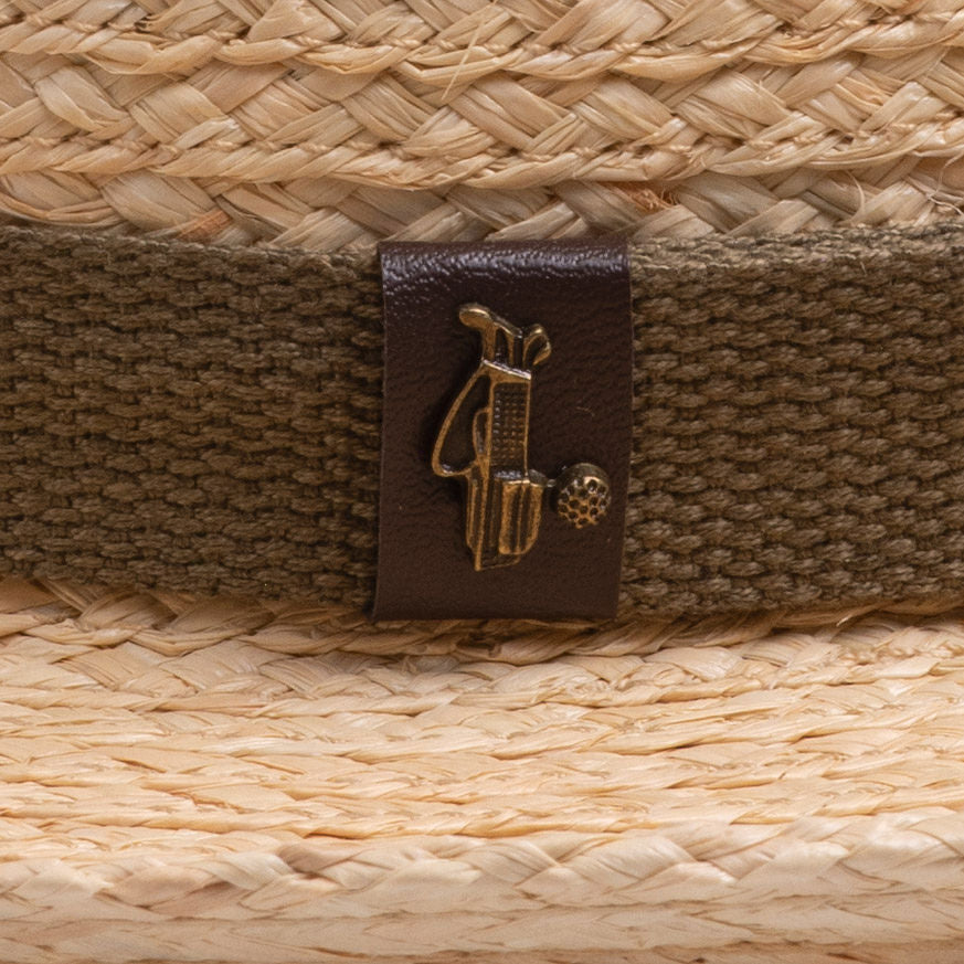 Close-up of a natural colored straw hat with a brown band and logo, featuring a golden colored golf club bag and golf ball ball pin on top of it