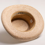 Underside of a natural colored straw hat with a golden colored sweatband on a light beige background