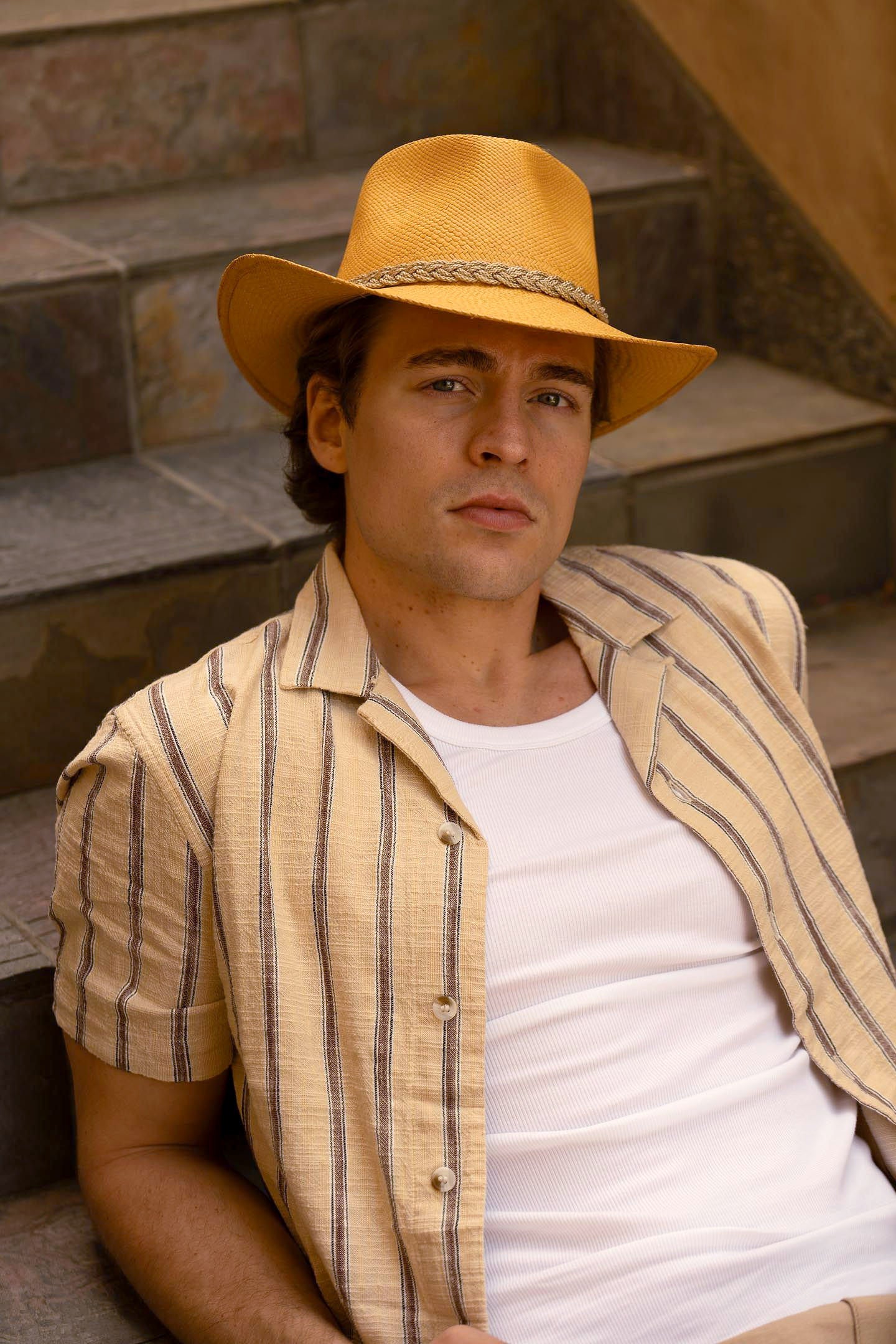 Man wearing a bright golden colored hat, striped shirt, and white undershirt, leaning back on gray stone steps