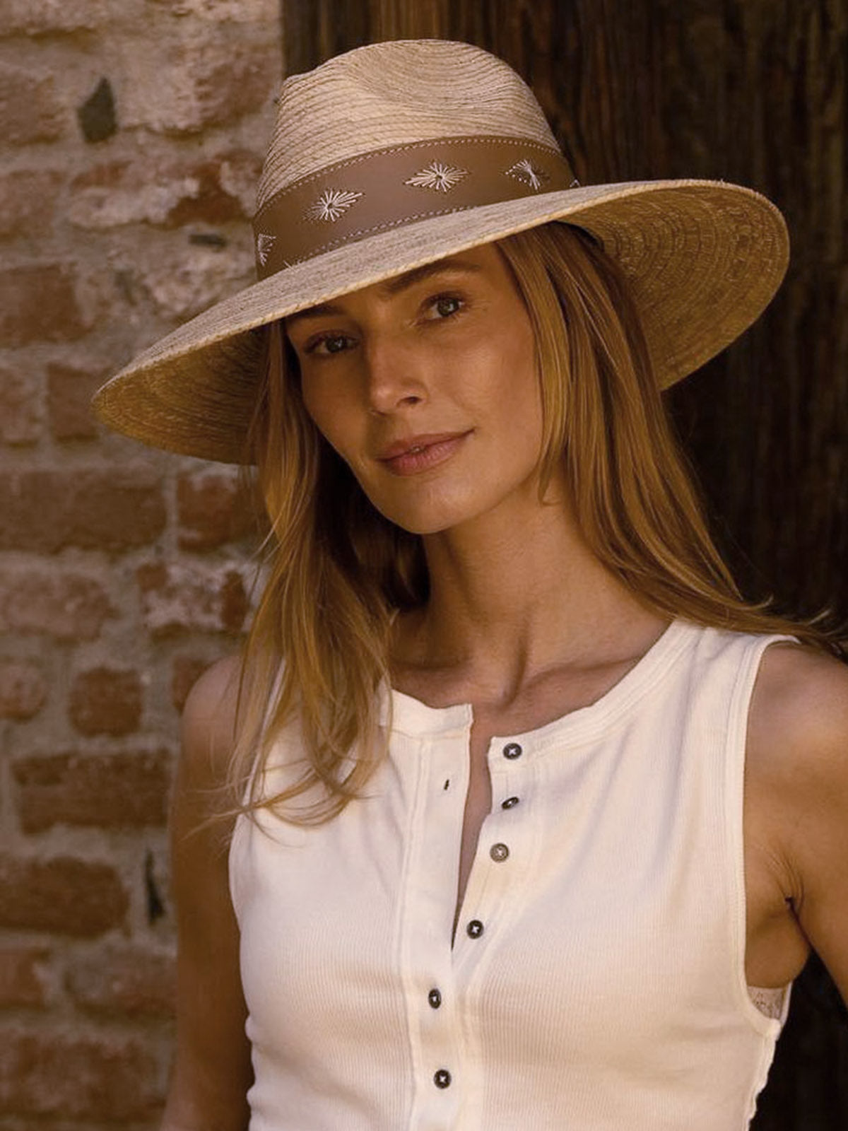 Woman wearing a wide-brimmed brown hat with a thick dark brown band standing near an entry way by a rustic brick wall