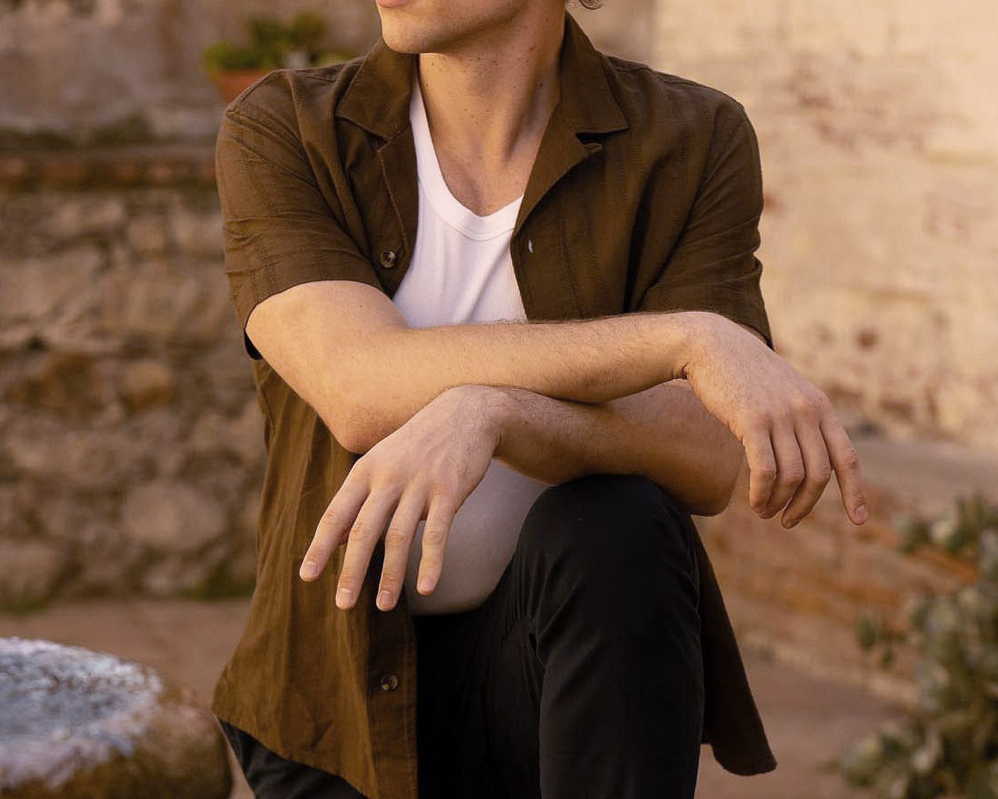 Man wearing a beige hat, brown shirt, white undershirt, and black pants, standing with one leg raised on a stone fountain with a stone wall background