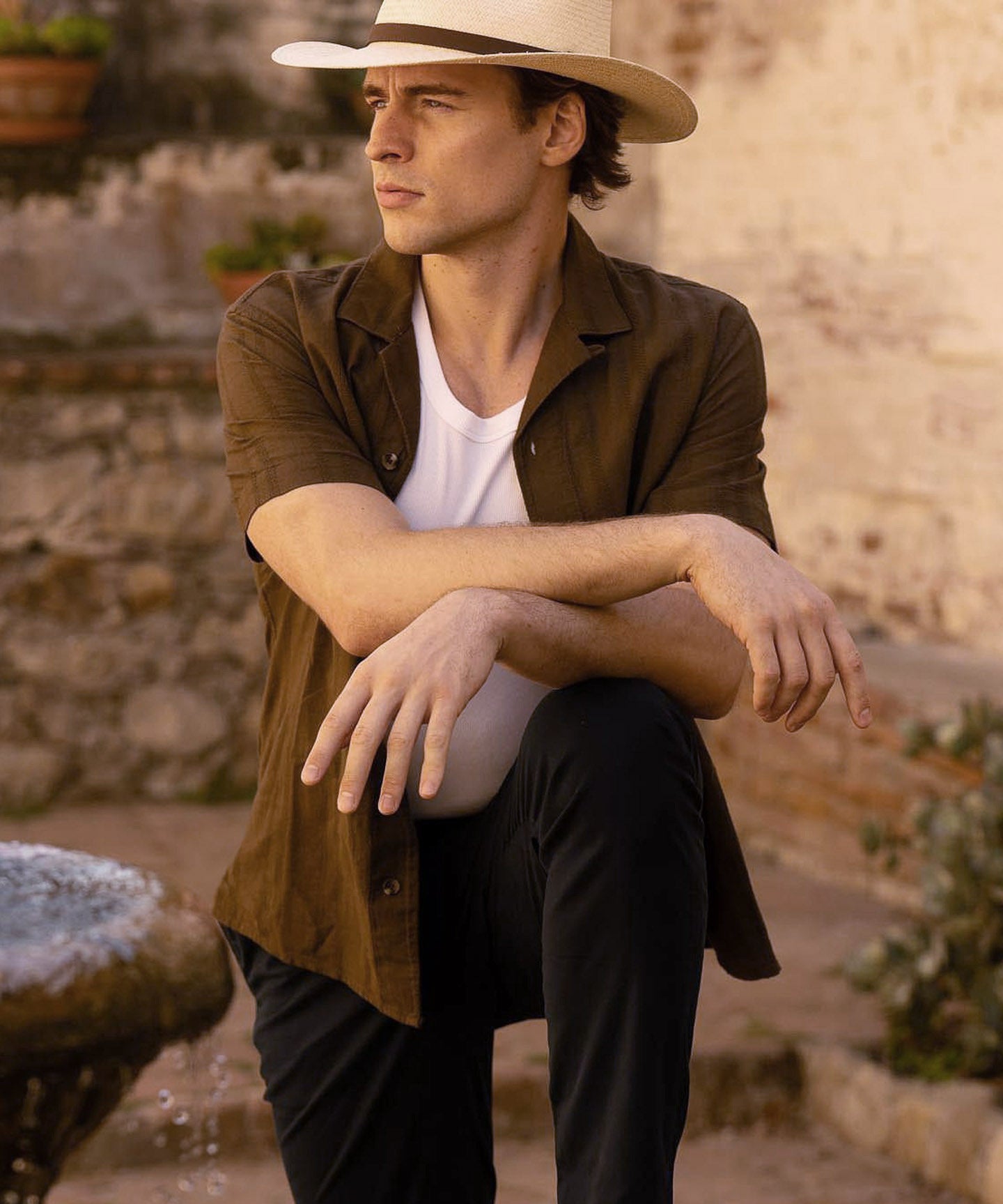 Man wearing a beige hat, brown shirt, white undershirt, and black pants, standing with one leg raised on a stone fountain with a stone wall background