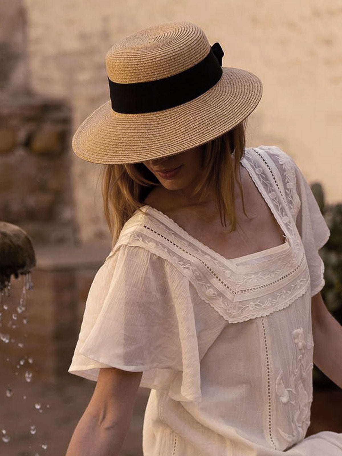 Woman in a yellow hat with thick black band and white dress playing with her arm outstretched with a water fountain while sitting in a garden