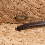 Close-up of a natural colored straw hat with a Scala logo pin and a brown leather strap going into a golden colored eyelet