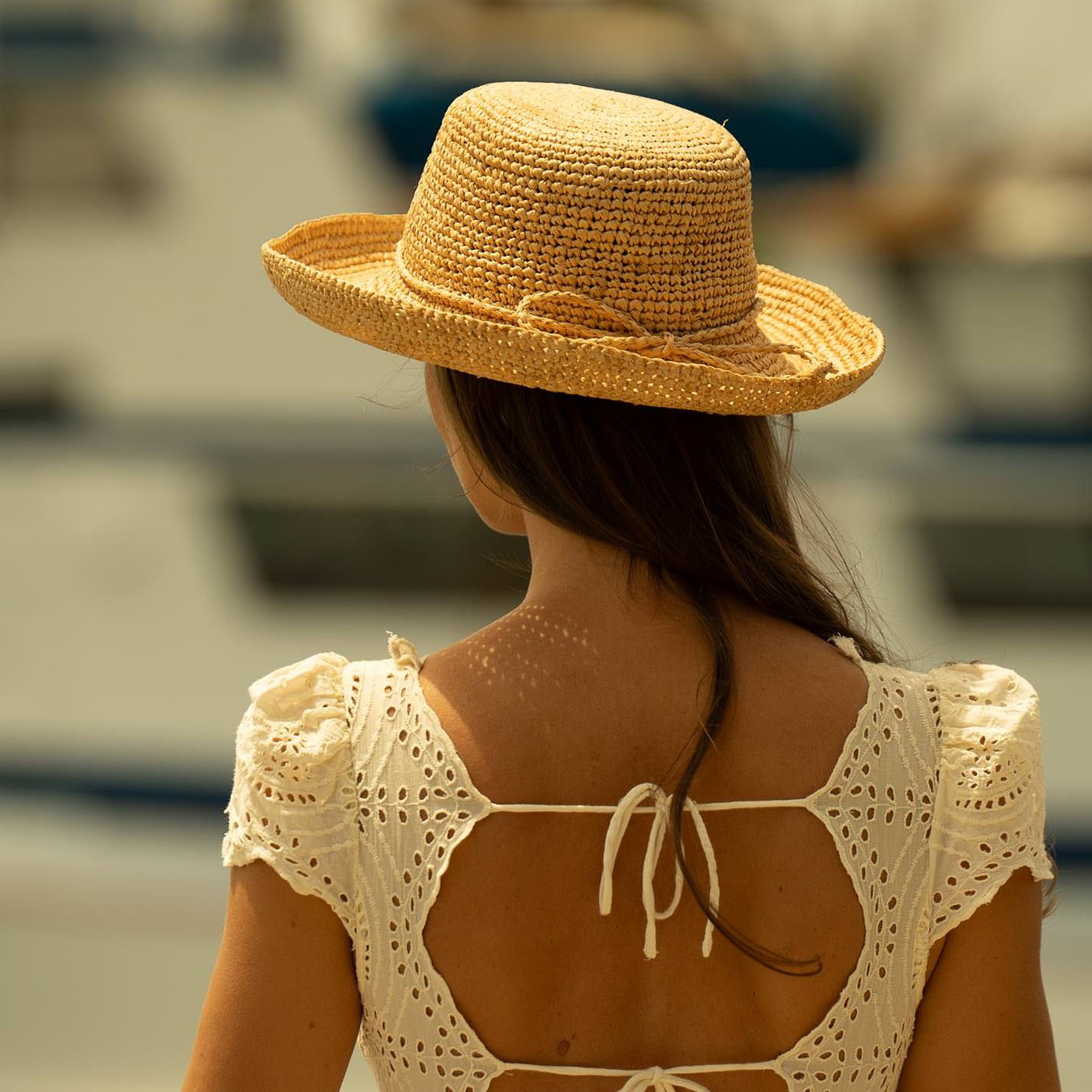 Woman wearing a natural colored straw hat and crochet top, facing away towards a blurred boat in the background