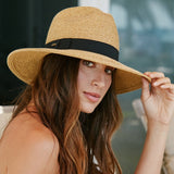 Woman wearing a toast colored straw hat with a black band and using her fingers to tilt the hat down a bit while outdoors