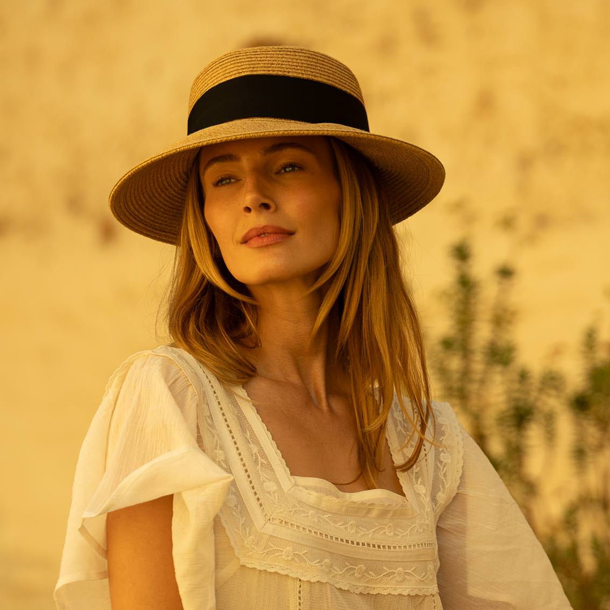 Woman wearing a tea colored straw hat with a thick black ribbon around it and a white blouse against a warm, blurred background