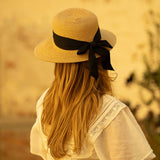 Woman wearing a tea colored straw hat with a thick black ribbon hanging off the back in a blurred natural setting