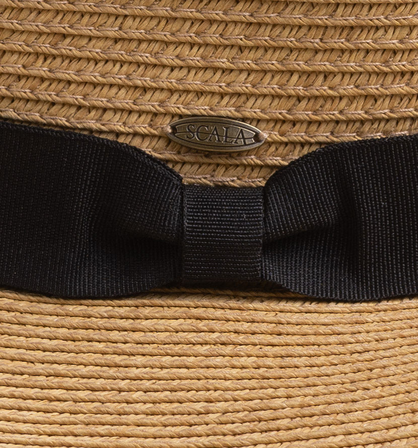 Close-up of a woven straw hat with a wide black ribbon bow and a small oval Scala brand pin at the center