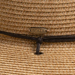 Close-up of a natural colored straw hat with a Scala logo pin, featuring a brown braided drawstring going into a gold eyelet