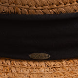 Close-up of a tea colored straw hat with a thick black ribbon and Scala logo pin on top of it