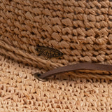 Close-up of a natural colored straw hat with a Scala logo pin and brown drawstring going into a golden colored eyelet