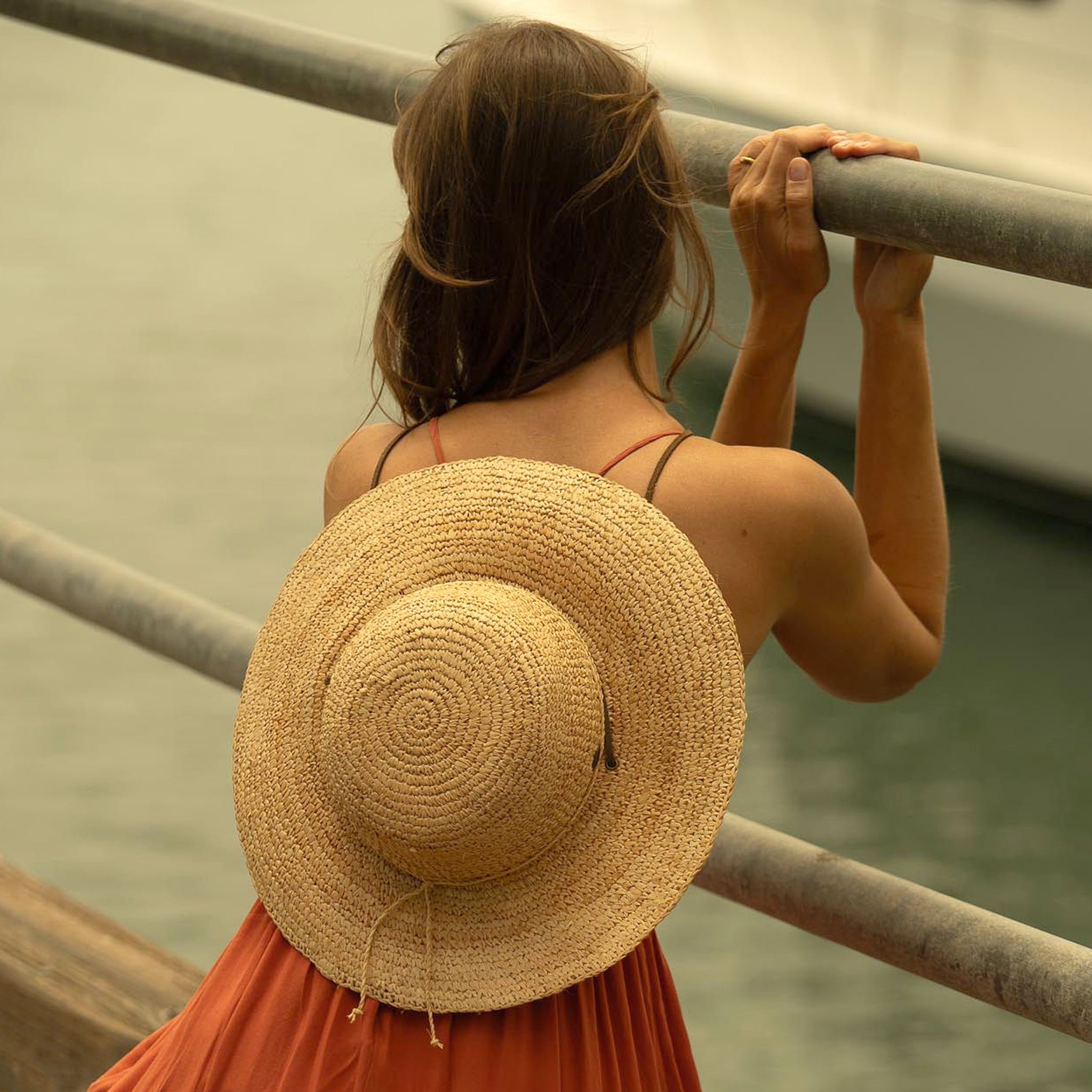 Woman using the drawstrings of her natural colored straw hat to hang off of her backside, wearing an orange dress and squatting down by a railing with a blurred river and boat in the background