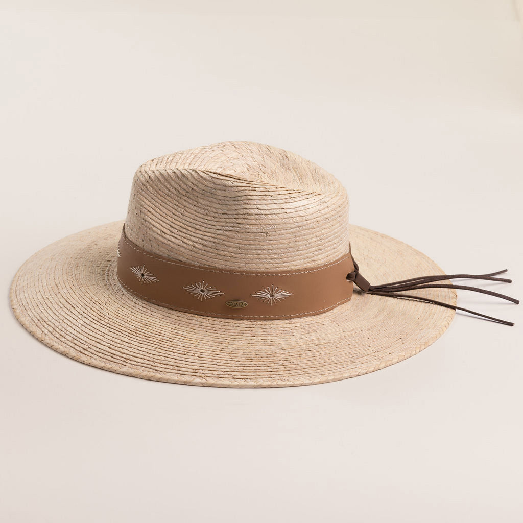 Natural colored straw hat with a brown band featuring white patterns and brown leather strings hanging off the back on a light beige background