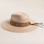 Natural colored straw hat with a brown band featuring white patterns and brown leather strings hanging off the back on a light beige background