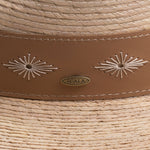 Close-up of a natural colored straw hat with decorative stitching and Scala logo pin on top of it