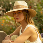 Woman wearing a natural colored straw hat with thick light brown band sitting on a bench in a garden setting