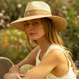 Woman wearing a natural colored straw hat with thick light brown band sitting on a bench in a garden setting