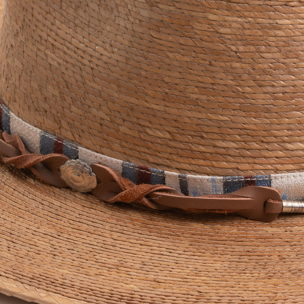 Close-up of a toast colored straw hat with a decorative band featuring leather and fabric elements