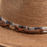 Close-up of a toast colored straw hat with a decorative band featuring leather and fabric elements