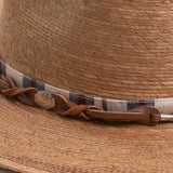 Close-up of a toast colored straw hat with a decorative band featuring leather and fabric elements