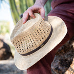 Man holding a natural colored straw hat on top of his red pants and knee with a cactus in the background