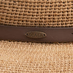 Close-up of a natural colored straw hat with a brown strap and Scala logo pin on top of it