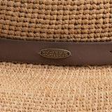 Close-up of a natural colored straw hat with a brown strap and Scala logo pin on top of it