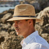 Man wearing a natural colored straw hat and white shirt standing against a rocky beachside
