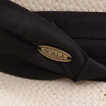 Close-up of a natural color straw hat with black band and Scala logo pin on top of it
