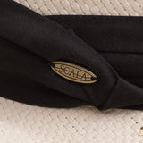 Close-up of a natural color straw hat with black band and Scala logo pin on top of it