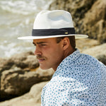 Man wearing a natural colored straw hat with a black band, sitting by a rocky shoreline