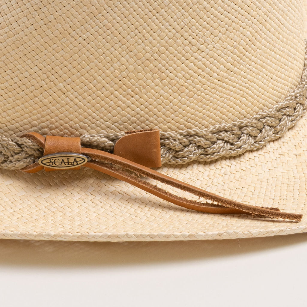 Close-up of a natural colored straw hat with sandy colored braided band and brown small ribbon with the Scala logo pin on top of it