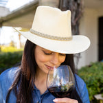 Woman wearing a natural colored hat and blue shirt, smelling a glass of red wine outdoors.