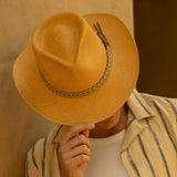 Man tipping his putty colored straw hat with a brown band down to cover his face against a beige background