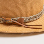 Close-up of a putty colored straw hat with sandy colored braided band and brown small ribbon with the Scala logo pin on top of it