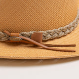 Close-up of a putty colored straw hat with sandy colored braided band and brown small ribbon with the Scala logo pin on top of it