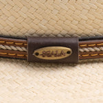 Close-up of a natural colored straw hat with a brown band and the Scala logo pin on top of it