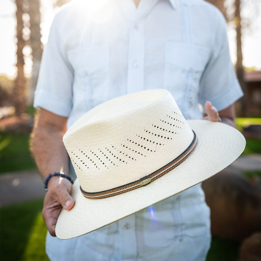 Man waring a shite shirt and light blue pants holding a natural colored straw hat with a blurred outdoor background