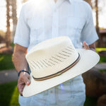 Man waring a shite shirt and light blue pants holding a natural colored straw hat with a blurred outdoor background