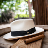 Bleach colored Panama hat with a thick black band and Scala logo pin resting on a wooden surface outdoors next to a cigar