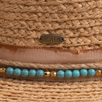 Close-up of a natural colored straw hat with a decorative band featuring turquoise beads and gold accents, with a Scala logo pin