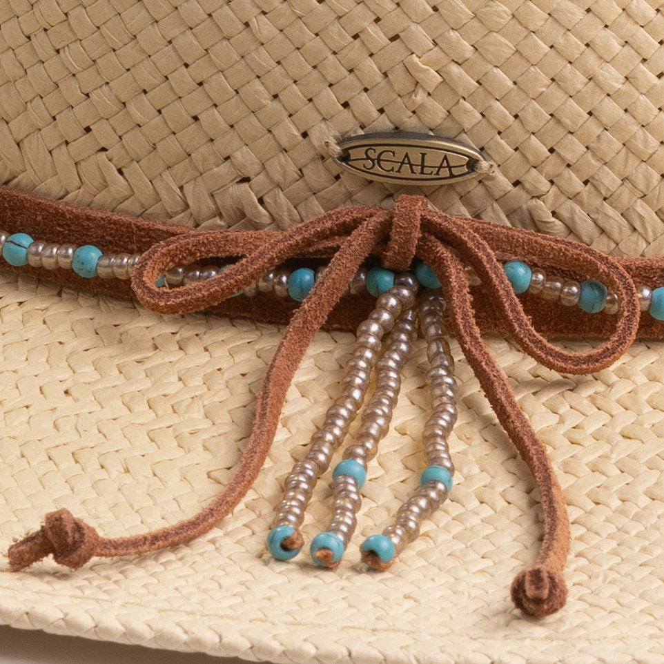 Close-up of a natural colored straw hat with decorative brown strings, turquoise and golden brown beads beads, and featuring the Scala logo pin
