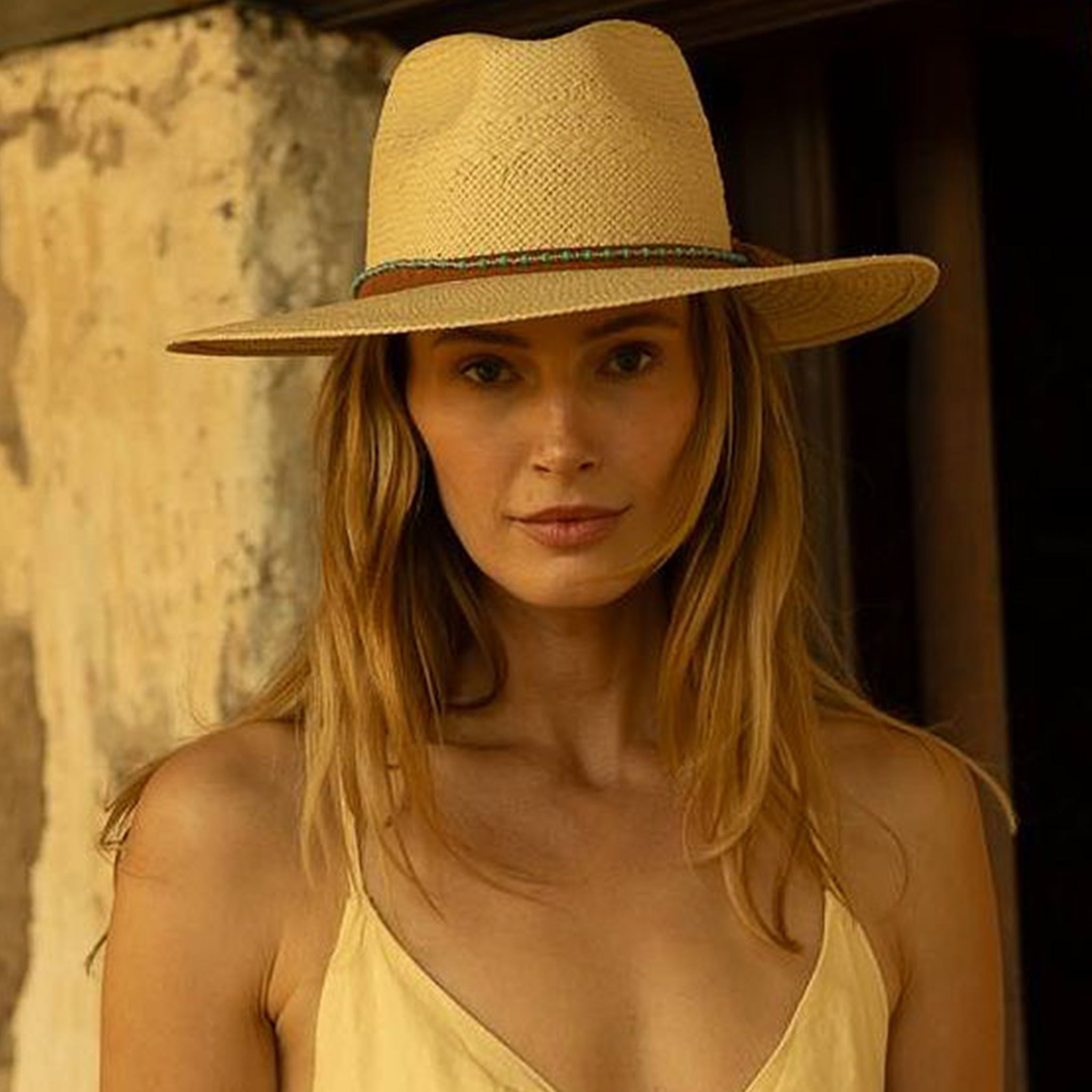 Woman wearing a natural colored straw hat and yellow top against a rustic wall.