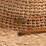 Close-up of a natural colored straw hat with the Scala logo pin and light brown band going into an brown eyelet