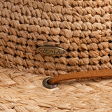Close-up of a natural colored straw hat with the Scala logo pin and light brown band going into an brown eyelet