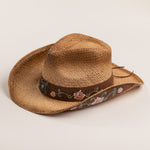 Tea colored straw cowboy-shaped hat with decorative floral pattern band and underbrim on a light beige background